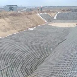 Skarpa wyłożona ażurowymi płytami betonowymi, zabezpieczająca wykop ziemny. Widoczne prace ziemne w tle i robotnicy w oddali.