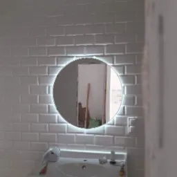 Białe, błyszczące płytki ułożone w cegiełkę na ścianie łazienki, z okrągłym lustrem z podświetleniem LED. Widoczne fragmenty umywalki i akcesoriów łazienkowych.