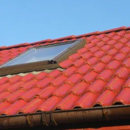 Czerwona dachówka ceramiczna z oknem dachowym i rynną w słoneczny dzień.