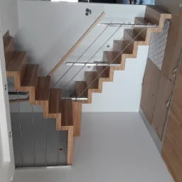 Ażurowe schody z jasnego drewna, z metalowymi relingami, zamontowane przy białej ścianie, widoczne zabezpieczenia podłogi.