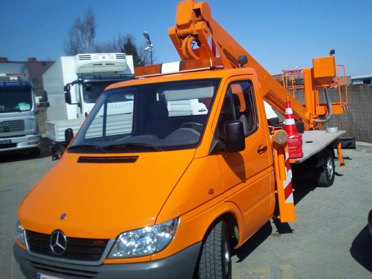 Pomarańczowy podnośnik koszowy na samochodzie Mercedes w Laskach, z widocznym stożkiem ostrzegawczym i elementami zabezpieczającymi na platformie, na tle innych pojazdów.