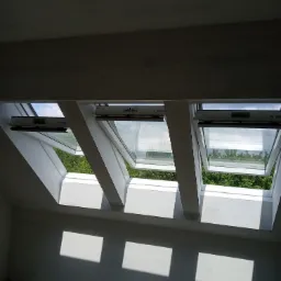 Trzy otwarte okna dachowe w jasnym, surowym wnętrzu, rzucające prostokątne cienie na ścianę, widok na zieleń na zewnątrz.