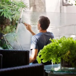 Mężczyzna czyści szklaną balustradę balkonu za pomocą ściągaczki, widoczne smugi wody i roślina doniczkowa na pierwszym planie.