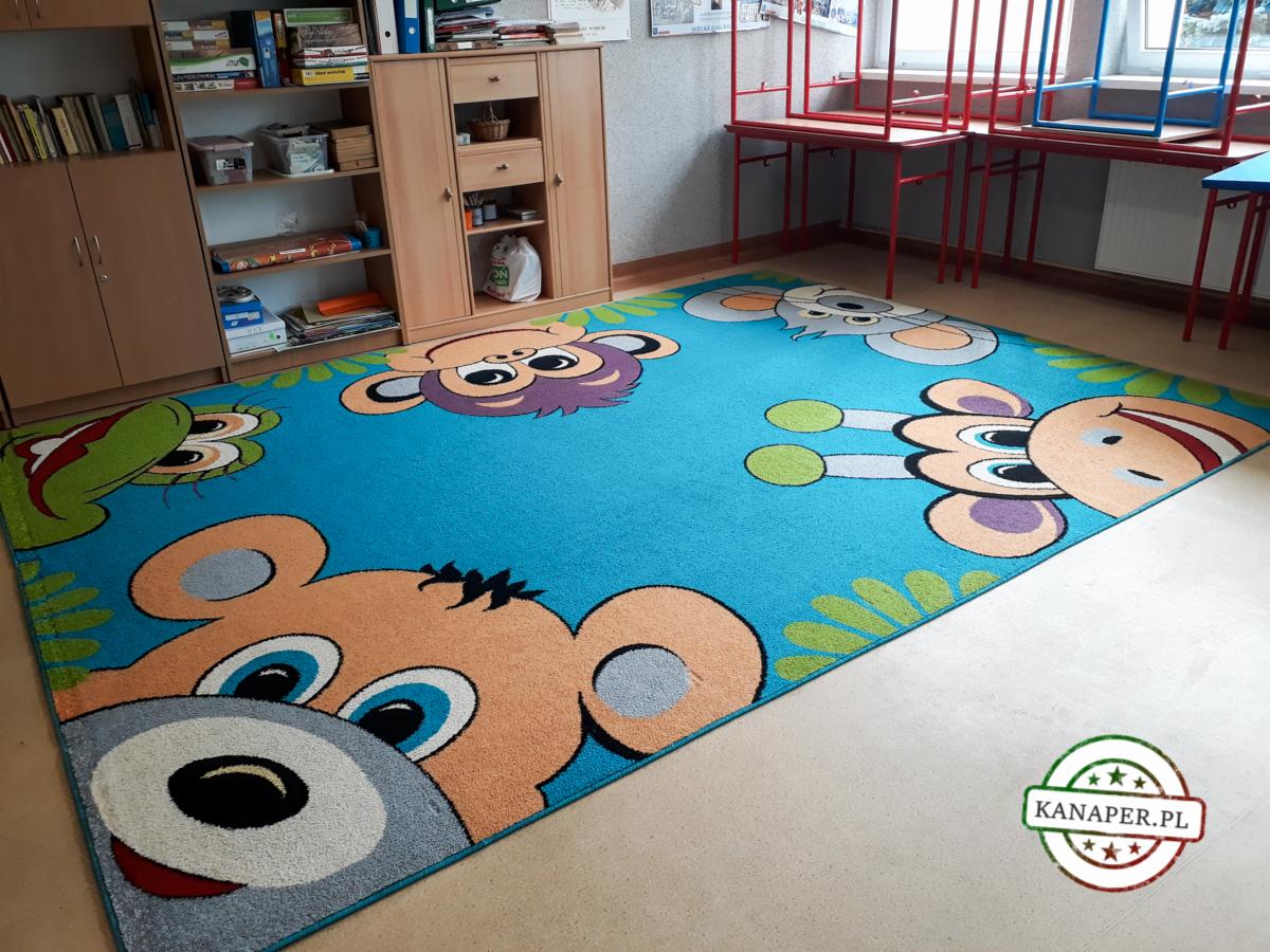 Niebieski dywan dziecięcy z motywami zwierząt (niedźwiedź, żyrafa, małpa, krokodyl) w pokoju, widoczne regały z książkami i zabawki w tle.