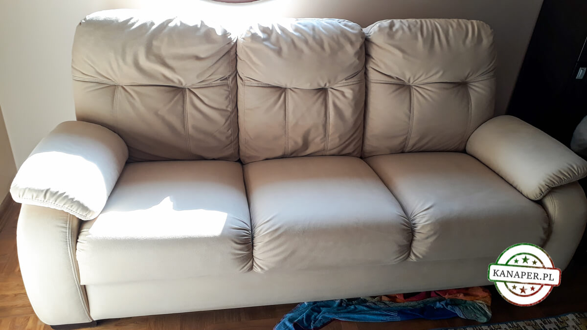 Jasna, trzyosobowa kanapa z poduszkami na oparciu, z widocznym logo 'kanaper.pl' w prawym dolnym rogu i częściowo widocznym dywanem pod spodem.