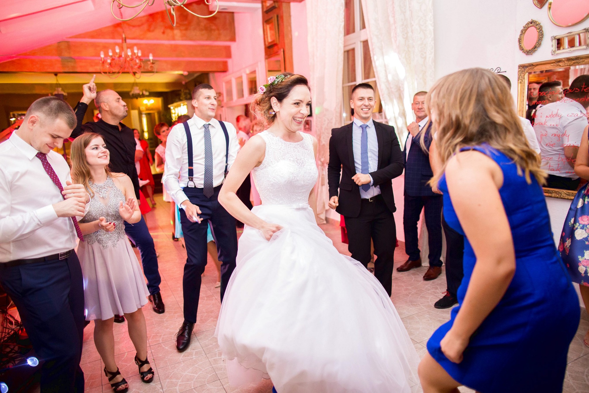 Uśmiechnięta panna młoda w białej sukni tańczy z gośćmi weselnymi na parkiecie, widoczne dekoracje sali i oświetlenie.