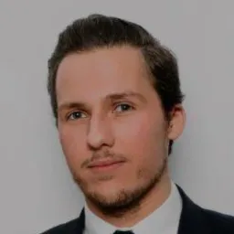 Portret młodego mężczyzny w ciemnym garniturze i krawacie na jasnoszarym tle. Uśmiech, krótka broda, profesjonalny wygląd.