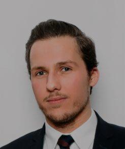 Portret młodego mężczyzny w ciemnym garniturze i krawacie na jasnoszarym tle. Uśmiech, krótka broda, profesjonalny wygląd.