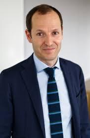 Portret biznesowy uśmiechniętego mężczyzny w ciemnej marynarce i błękitnym krawacie w paski, na jasnym tle. Dobre oświetlenie, widoczny profesjonalizm.