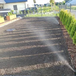 Przygotowany grunt pod trawnik, nawadniany zraszaczy, obrzeża z kamieni i kory, w tle dom z grafitowym dachem i tujami.