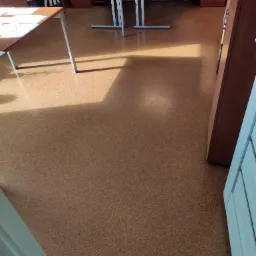 Widok na brązową podłogę w biurze z widocznymi biurkami i szafkami, oświetloną przez promienie słoneczne wpadające przez okno.