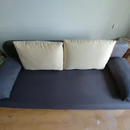 Szara kanapa z dwoma kremowymi poduszkami na drewnianej podłodze, widoczny fragment ściany z gniazdkiem elektrycznym.