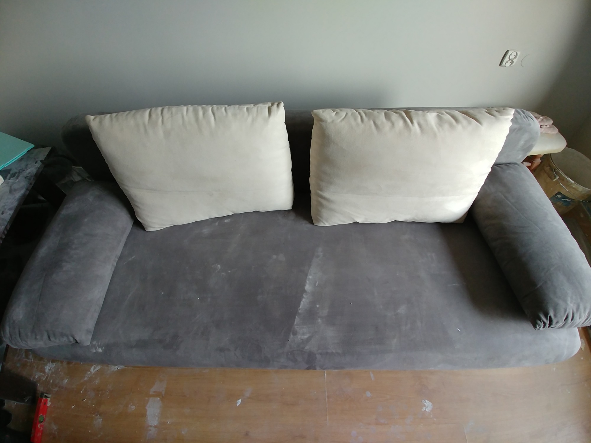 Szara kanapa z dwoma jasnymi poduszkami na tle ściany i drewnianej podłogi z widocznymi śladami po remoncie.