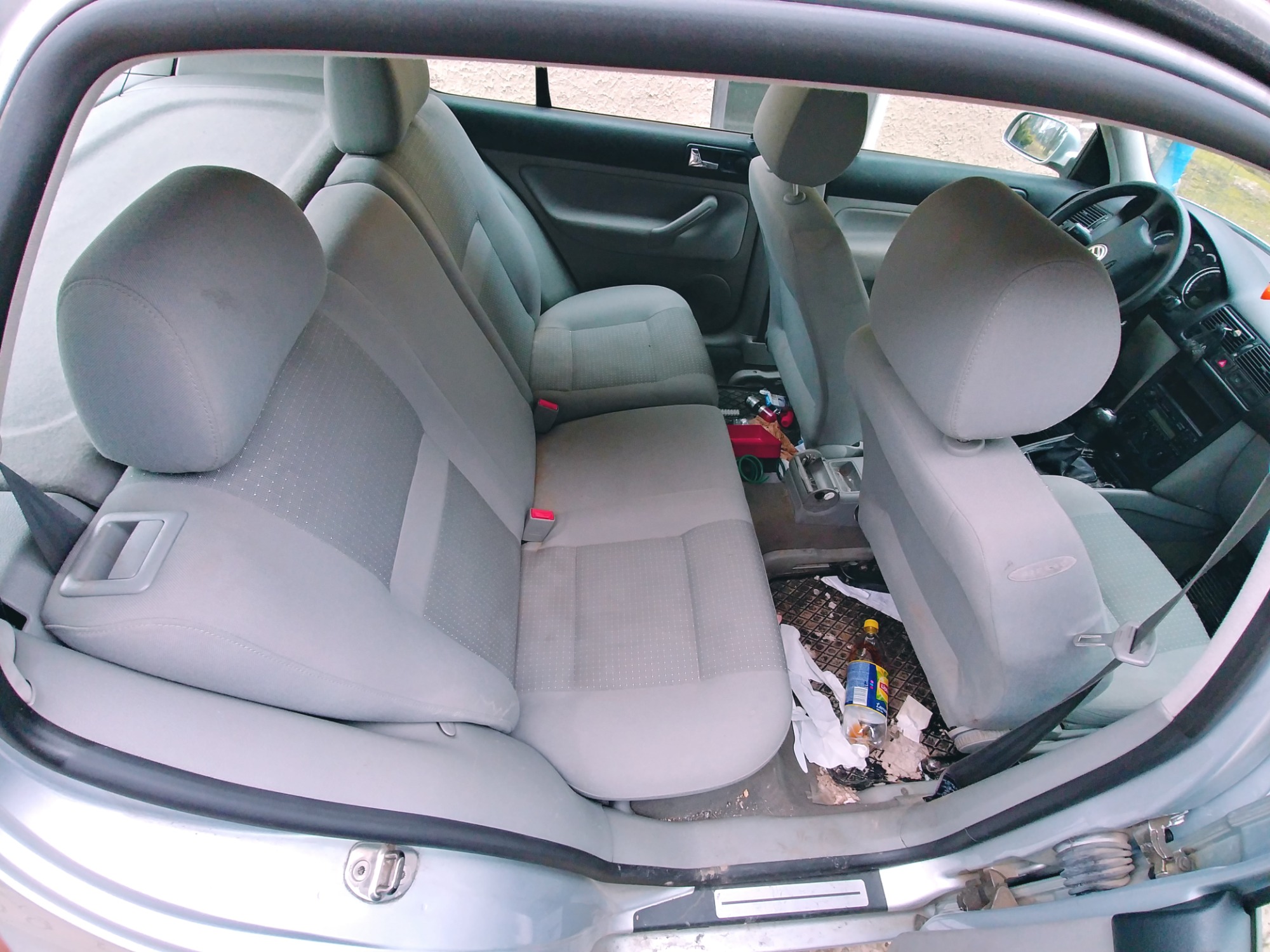 Wnętrze samochodu z szarą tapicerką, widoczne tylne siedzenia z zapiętymi pasami, na podłodze śmieci, butelka i rękawiczki, perspektywa z zewnątrz przez otwarte drzwi.