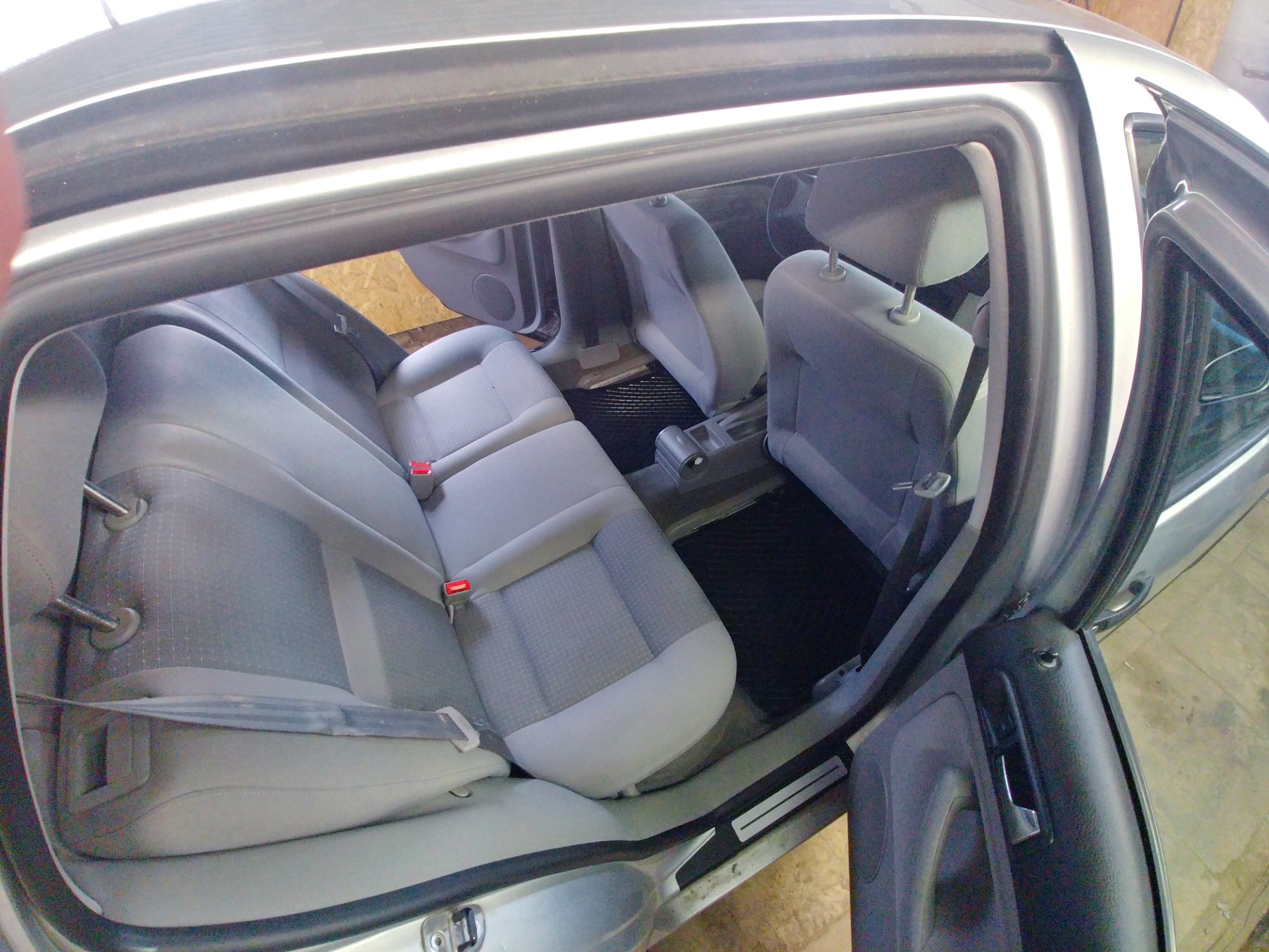 Wnętrze samochodu z otwartymi drzwiami, widoczne szare tapicerowane siedzenia z czerwonymi klamrami pasów bezpieczeństwa i czarne dywaniki, uchwycone z perspektywy zewnętrznej.