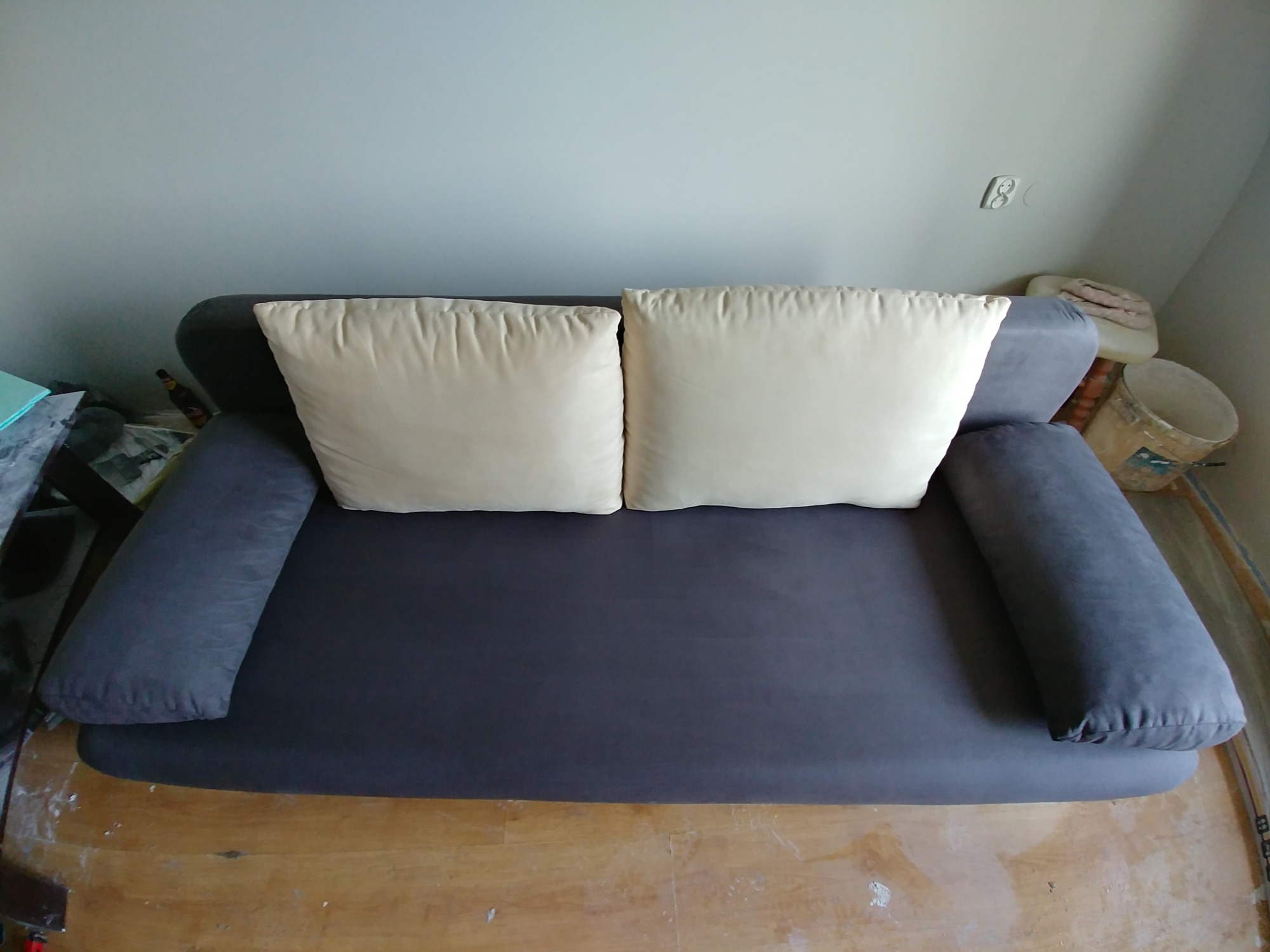 Szara kanapa z dwoma kremowymi poduszkami na drewnianej podłodze, widoczny fragment ściany z gniazdkiem elektrycznym.