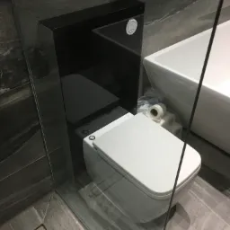 Nowoczesna toaleta wisząca z czarnym stelażem podtynkowym i przyciskiem spłukującym, obok wanna i zapas rolek papieru toaletowego, całość za szklaną ścianką kabiny prysznicowej, szare płytki...