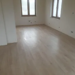 Jasne panele podłogowe w pokoju z dwoma oknami w drewnianych ramach, ściany w kolorze złamanej bieli, widoczne ślady po instalacji elektrycznej.