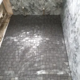 Świeżo ułożona mozaika w odcieniach szarości na podłodze i ścianach łazienki, widoczne rury instalacyjne i niebieskie wiadro z akcesoriami glazurniczymi w rogu.