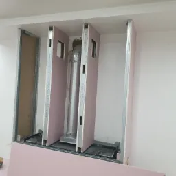 Konstrukcja z płyt kartonowo-gipsowych z metalowym stelażem i rurą wentylacyjną wewnątrz, widoczne narzędzia i materiały budowlane w pomieszczeniu.