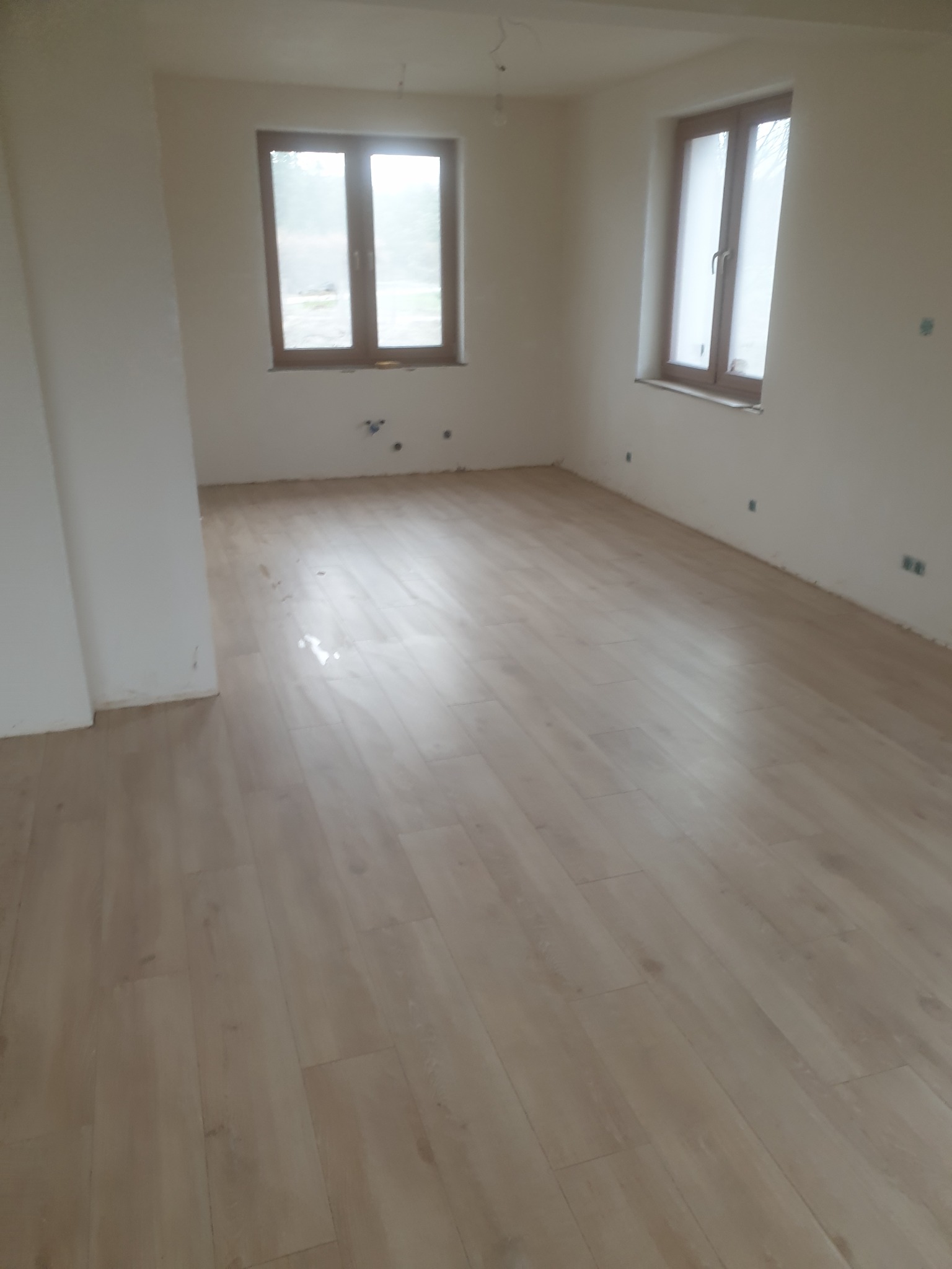 Jasne panele podłogowe w pokoju z dwoma oknami w drewnianych ramach, ściany w kolorze złamanej bieli, widoczne ślady po instalacji elektrycznej.