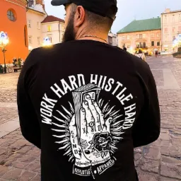 Mężczyzna w czarnej koszulce z długim rękawem, na której plecach widnieje biały nadruk z motywem dłoni trzymającej plik banknotów i napisem 'WORK HARD HUSTLE HARD GRIND', na tle brukowanego placu...