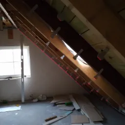 Poddasze w trakcie ocieplania z widoczną konstrukcją drewnianą, laserowo wyznaczonym poziomem, oknem i częściowo ułożoną izolacją.