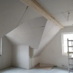 Poddasze z zamontowanymi płytami gipsowo-kartonowymi, widoczne drewniane belki stropowe, okno i drabina w jasnym, surowym wnętrzu.