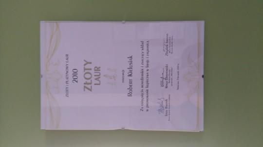 Dyplom 'Złoty Laur' dla Roberta Kielusiaka za zasługi w rozwoju kupiectwa, wiszący na zielonej ścianie.