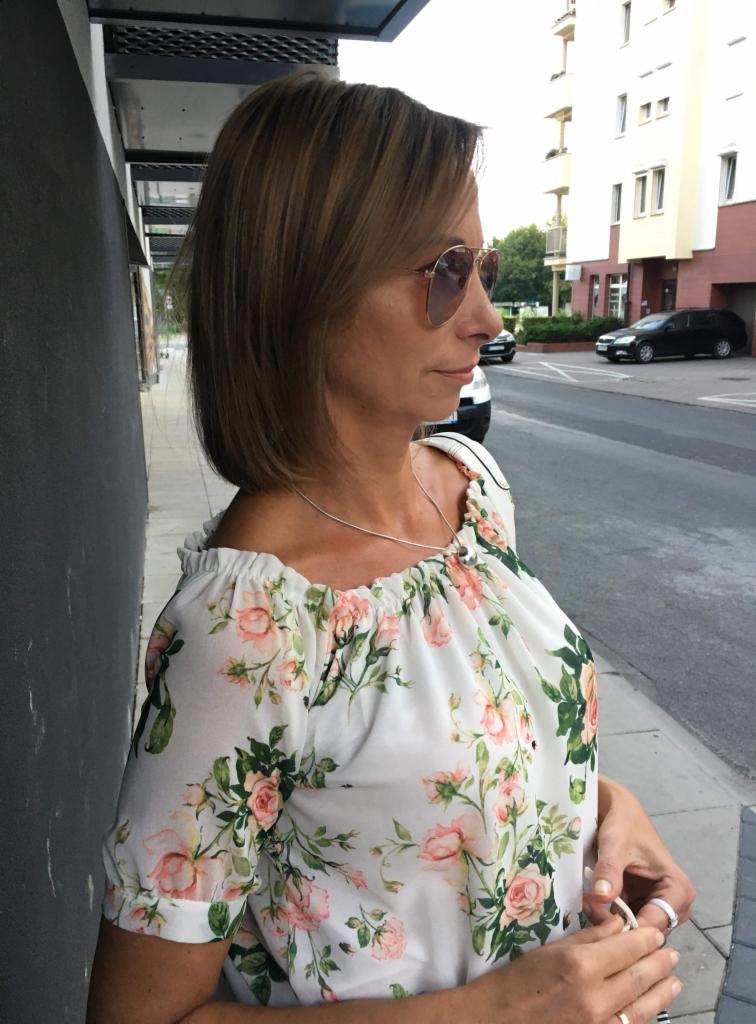 Portret kobiety w okularach przeciwsłonecznych, w bluzce w kwiaty, stojącej na tle miejskiej ulicy i budynku. Ujęcie z profilu.