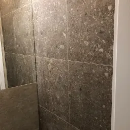 Ściana łazienki wyłożona dużymi, brązowo-szarymi płytkami imitującymi kamień z wyraźnymi, jasnymi inkluzjami i fugami w kolorze zbliżonym do płytek, widoczny fragment białej ościeżnicy drzwi.