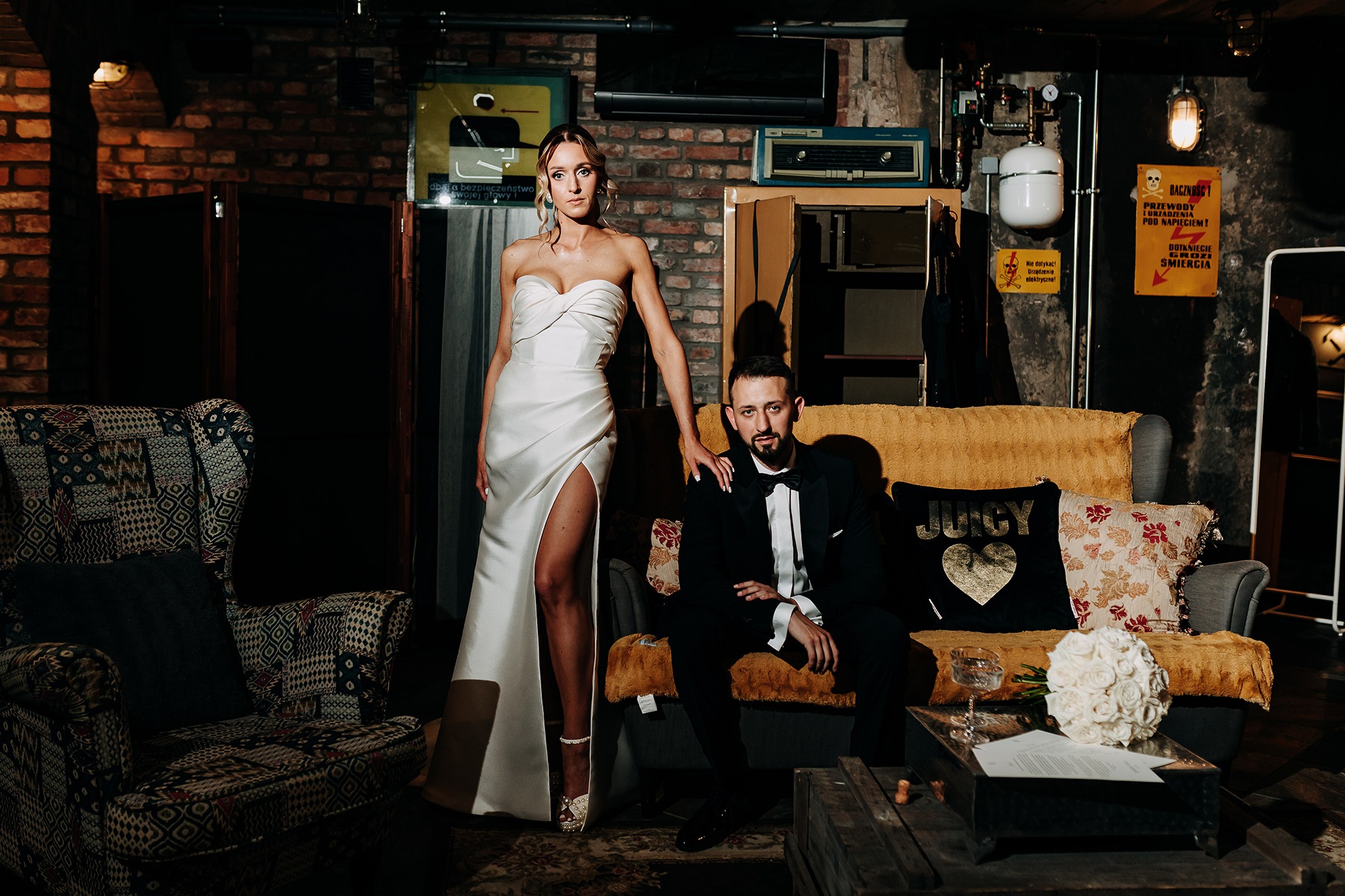 Elegancka sesja ślubna w loftowym wnętrzu: panna młoda w białej sukni z rozcięciem, pan młody w smokingu, bukiet róż, surowa cegła i industrialne detale.