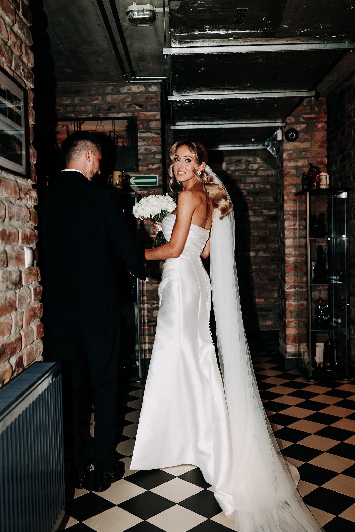 Panna młoda w białej sukni spogląda przez ramię, trzymając bukiet białych róż. Obok niej stoi pan młody w czarnym garniturze. W tle ceglana ściana i czarno-biała podłoga.