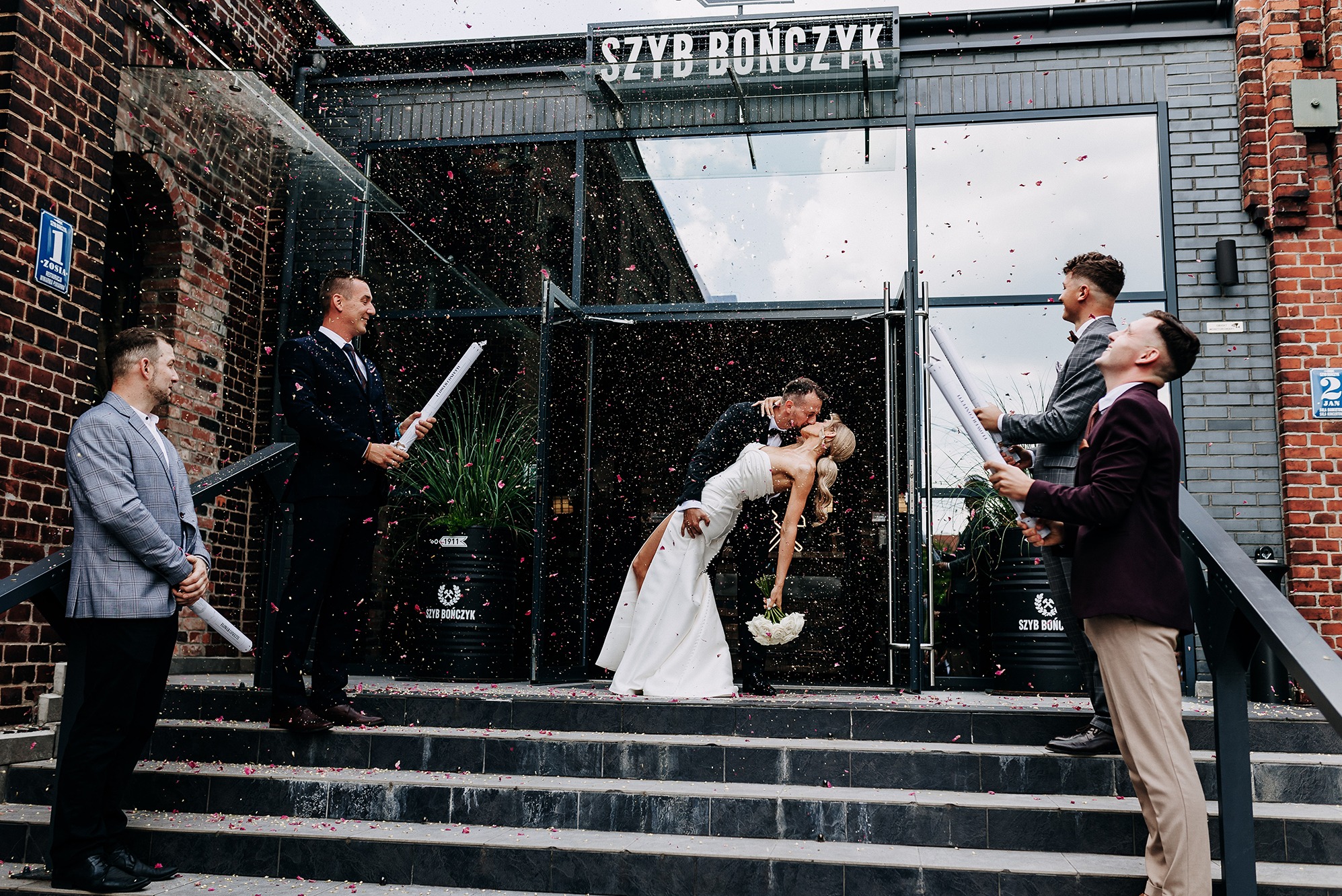 Pocałunek pary młodej na schodach 'Szybu Bończyk' w deszczu konfetti, otoczonej przez mężczyzn z tubami konfetti. Ujęcie z dołu, podkreślające radość chwili.