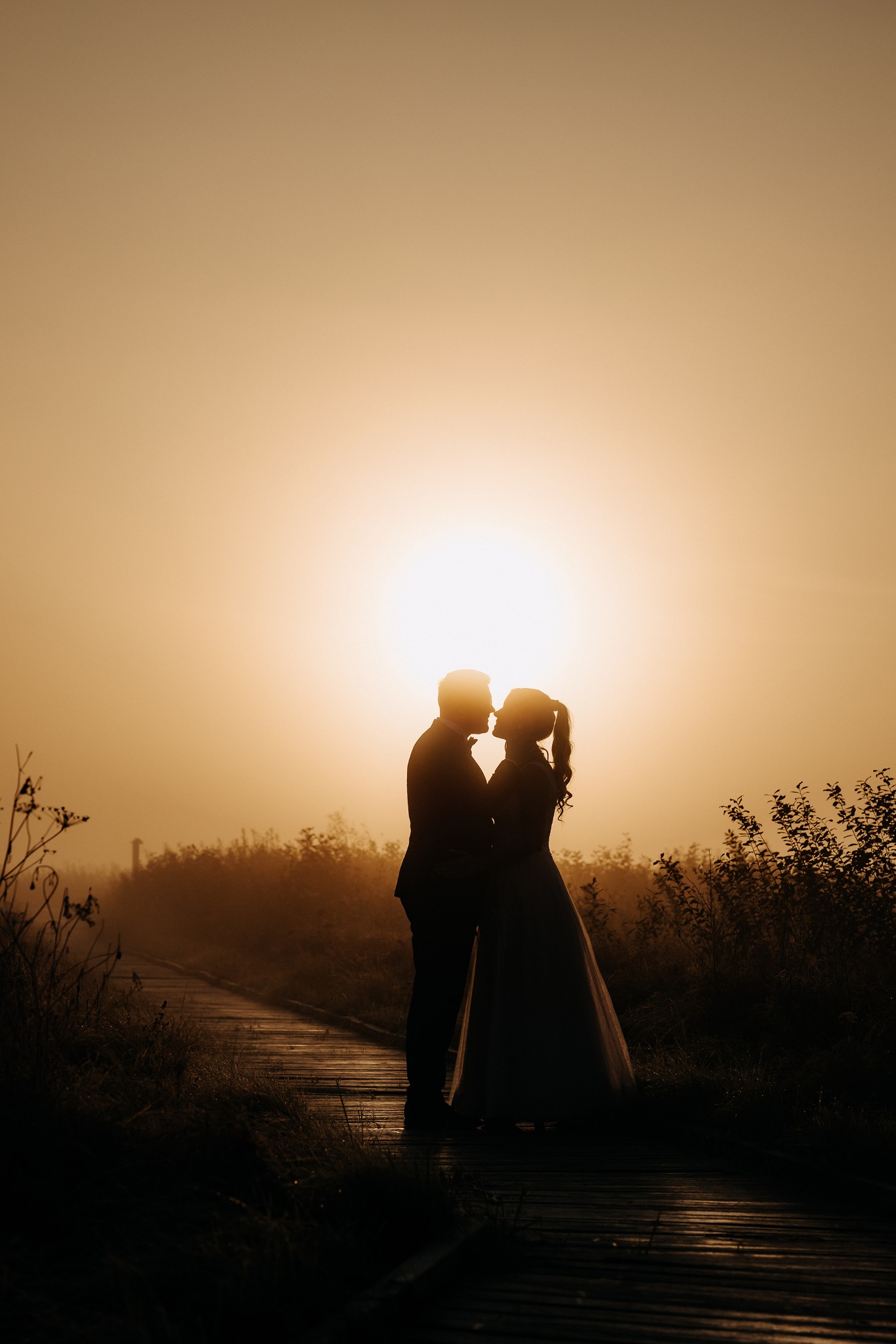 Sylwetki całującej się pary młodej na tle wschodzącego słońca, na drewnianej ścieżce w plenerze. Romantyczna atmosfera, ciepłe światło, delikatna mgła.