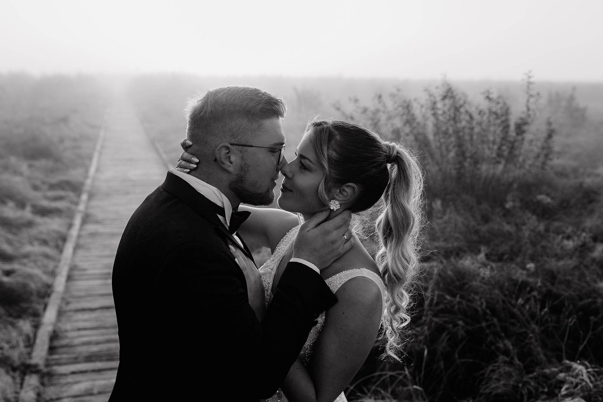 Czarno-białe zbliżenie całującej się pary młodej na tle mglistej drewnianej ścieżki. Pan młody w okularach i smokingu, panna młoda z upiętymi włosami i delikatną biżuterią.