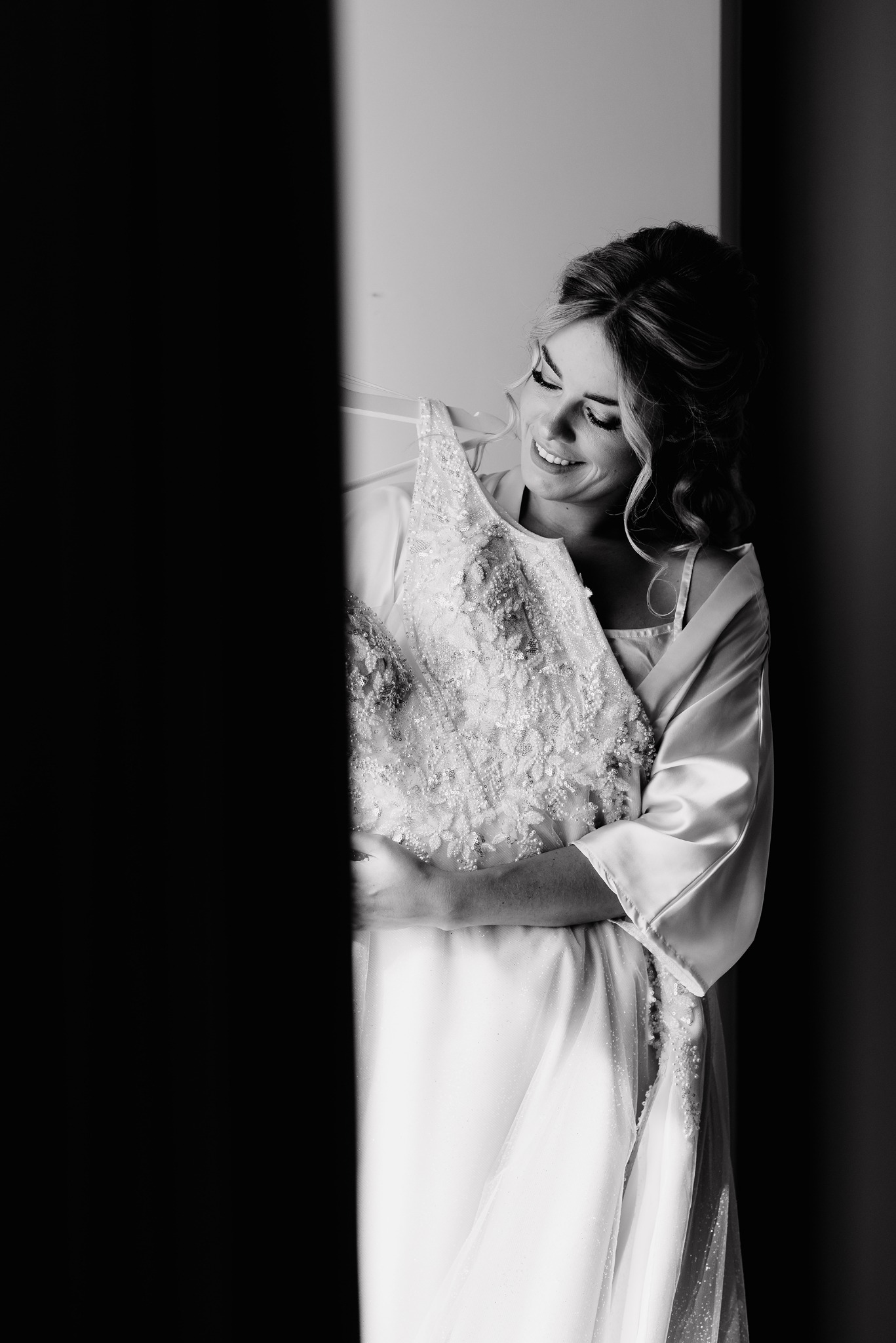 Uśmiechnięta panna młoda w satynowym szlafroku, trzymająca zdobioną suknię ślubną w czerni i bieli, z delikatnym światłem padającym z boku.