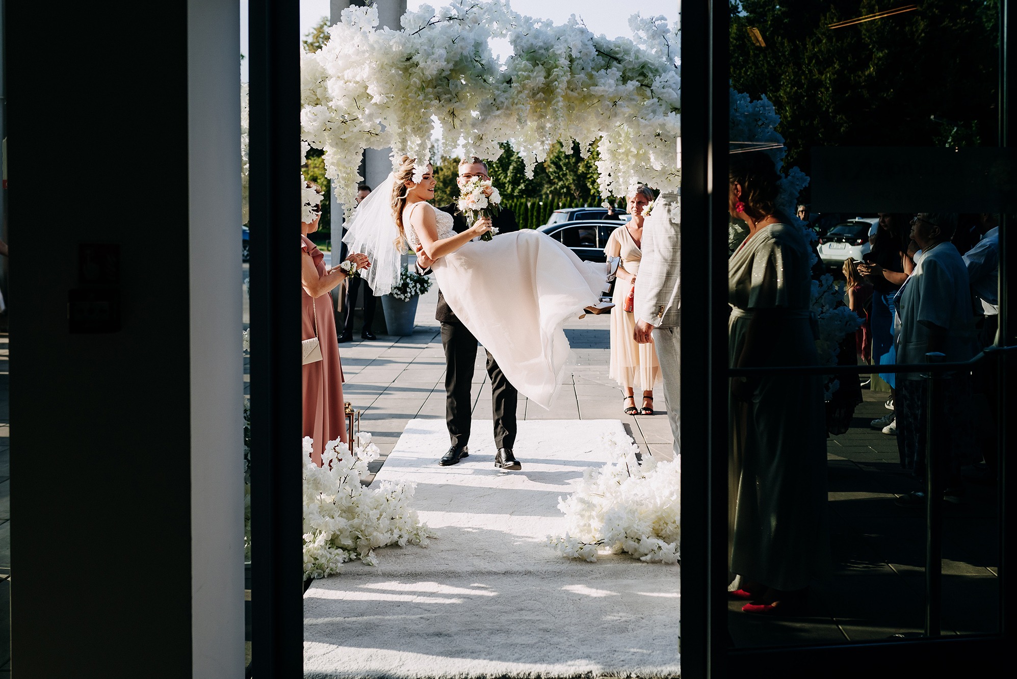 Pan młody trzyma pannę młodą na rękach pod kwiatowym łukiem, goście w tle, ujęcie zza szklanych drzwi, ciepłe światło.