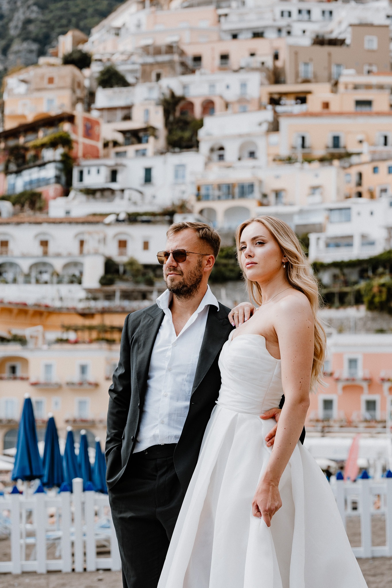 Elegancka para młoda w Positano: on w garniturze, ona w białej sukni, na tle malowniczych, kolorowych budynków i błękitnych parasoli plażowych.