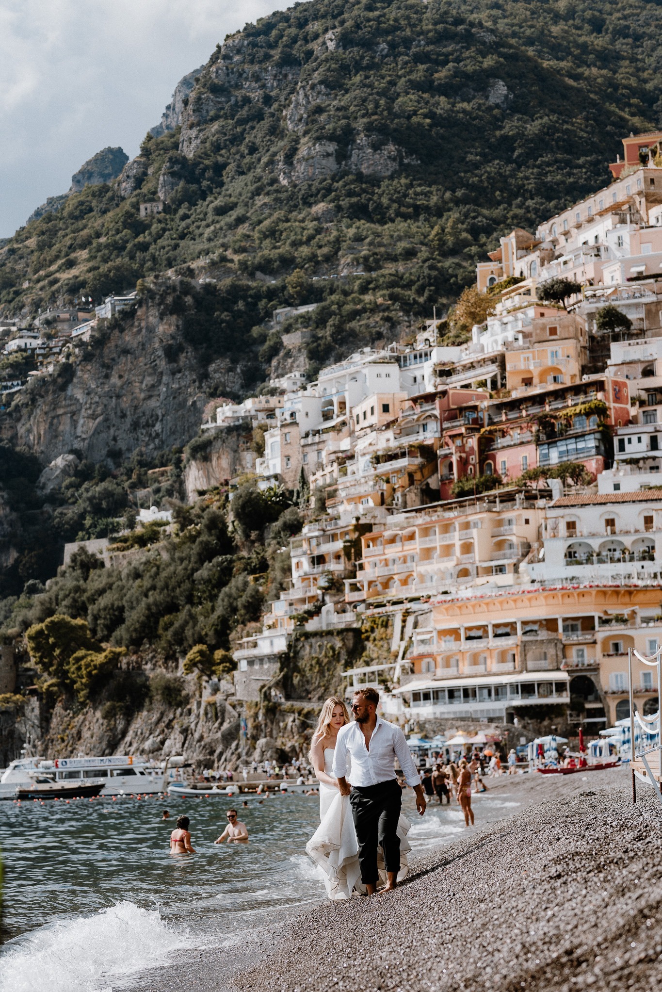 Para młoda spaceruje brzegiem morza w Positano, Włochy. W tle malownicze, kolorowe budynki na zboczu góry i błękitne morze. Ujęcie z dołu, podkreślające krajobraz.