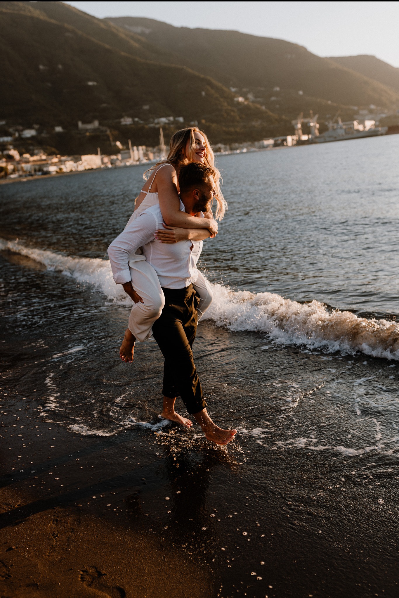 Zakochana para na plaży o zachodzie słońca. Kobieta na barana u mężczyzny, oboje boso w wodzie. W tle góry i zabudowania.