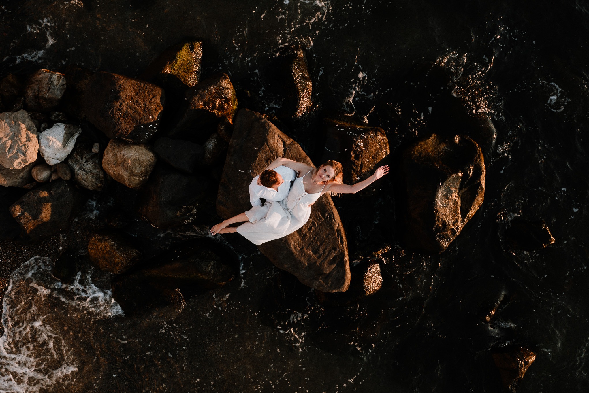 Para młoda w bieli, leżąca na skale wśród fal morskich, uchwycona z lotu ptaka w ciepłym świetle zachodzącego słońca. Romantyczna sesja na tle ciemnej wody.