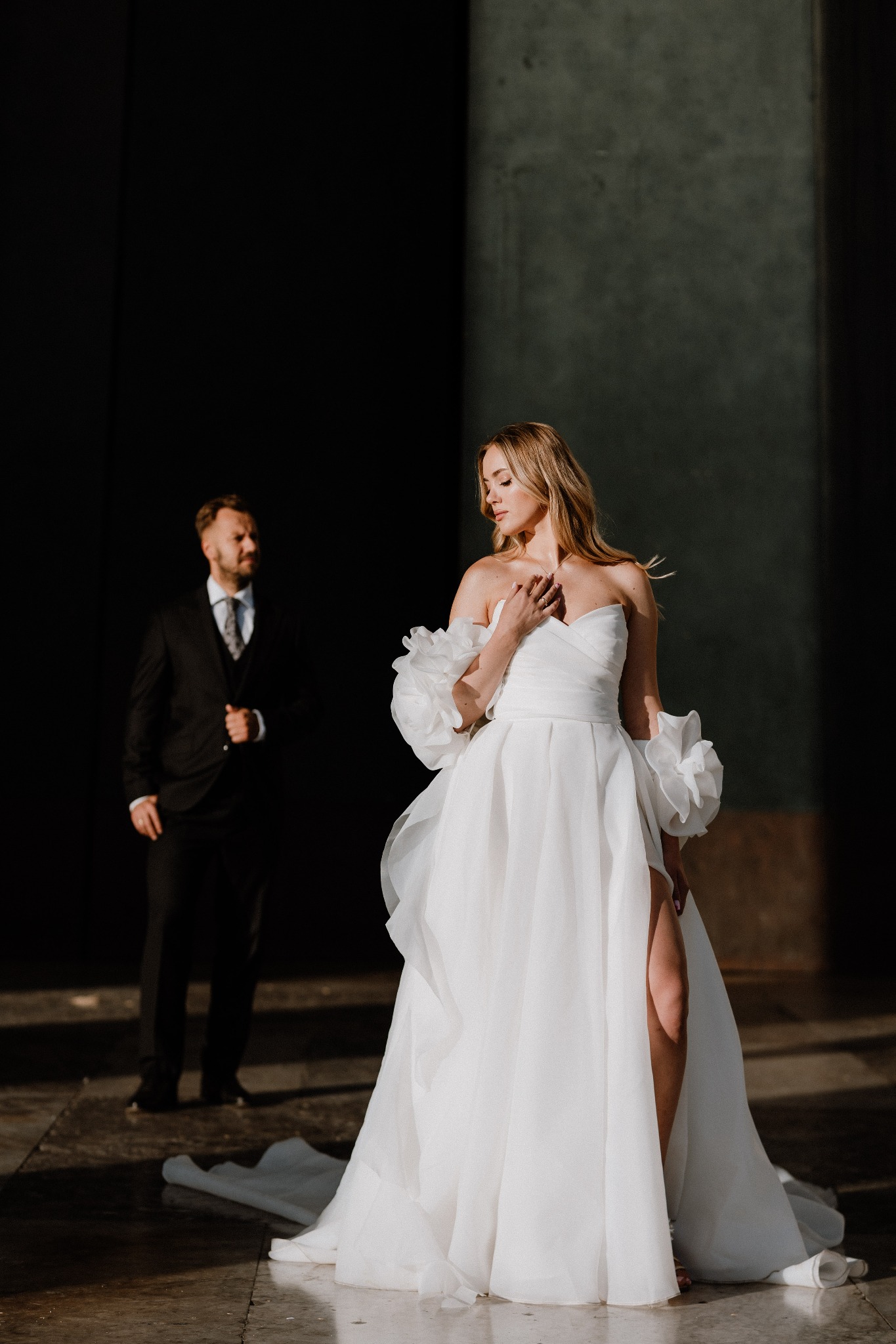 Elegancka sesja ślubna: panna młoda w białej sukni z rozcięciem, z falbanami na ramionach, pozuje na tle kolumn, w tle rozmyty pan młody w garniturze.
