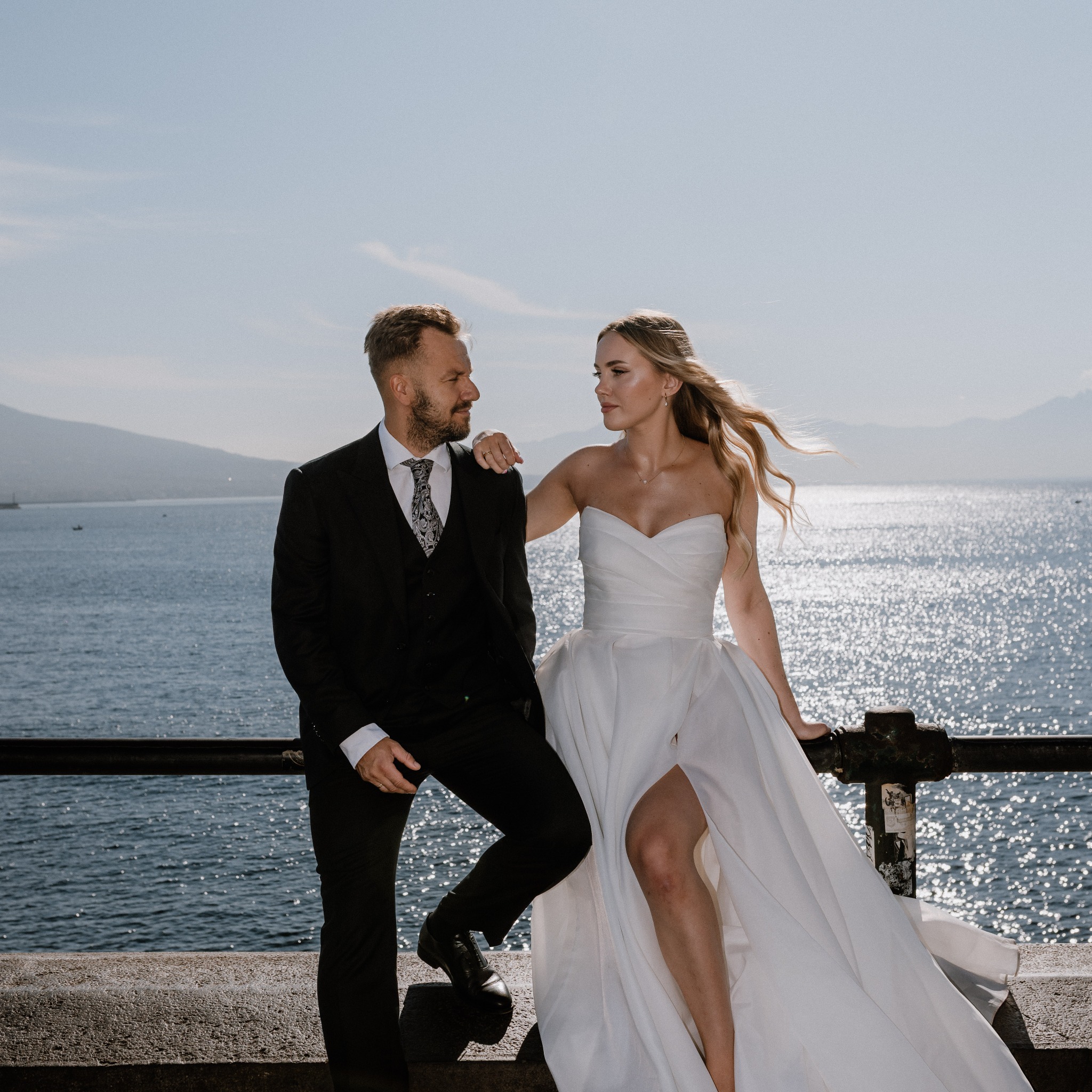 Elegancka para młoda w plenerze nad morzem. Panna młoda w białej sukni z rozcięciem, pan młody w czarnym garniturze. W tle błękitne morze i góry.