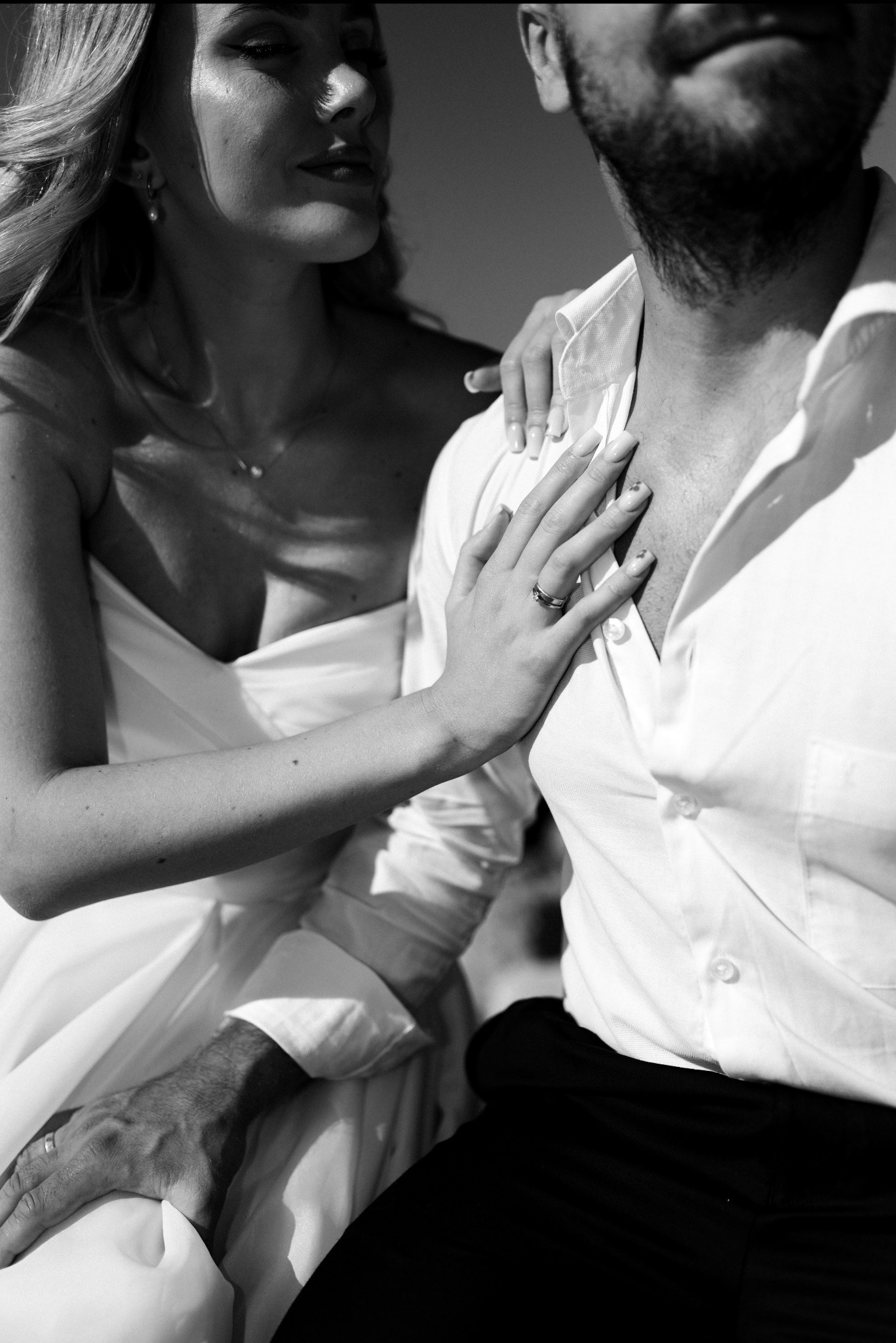 Czarno-białe zbliżenie pary: kobieta w sukni ślubnej opiera dłoń z obrączką na piersi mężczyzny w białej koszuli. Delikatne światło podkreśla detale.