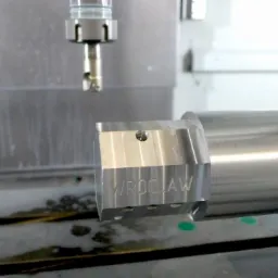 Metalowy detal z napisem 'WROCLAW' obrabiany na tokarce CNC, widoczny frez i uchwyt mocujący