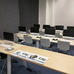 Sala komputerowa z rzędami stanowisk wyposażonych w komputery typu All-in-One, na biurkach leżą materiały szkoleniowe.