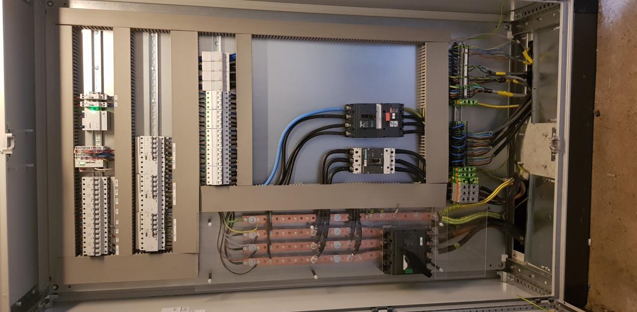 Otwarta szafa sterownicza z widocznymi elementami instalacji elektrycznej: wyłączniki, listwy zaciskowe, przewody w kolorach czarnym, niebieskim i żółto-zielonym, ułożone wewnątrz metalowej obudowy.