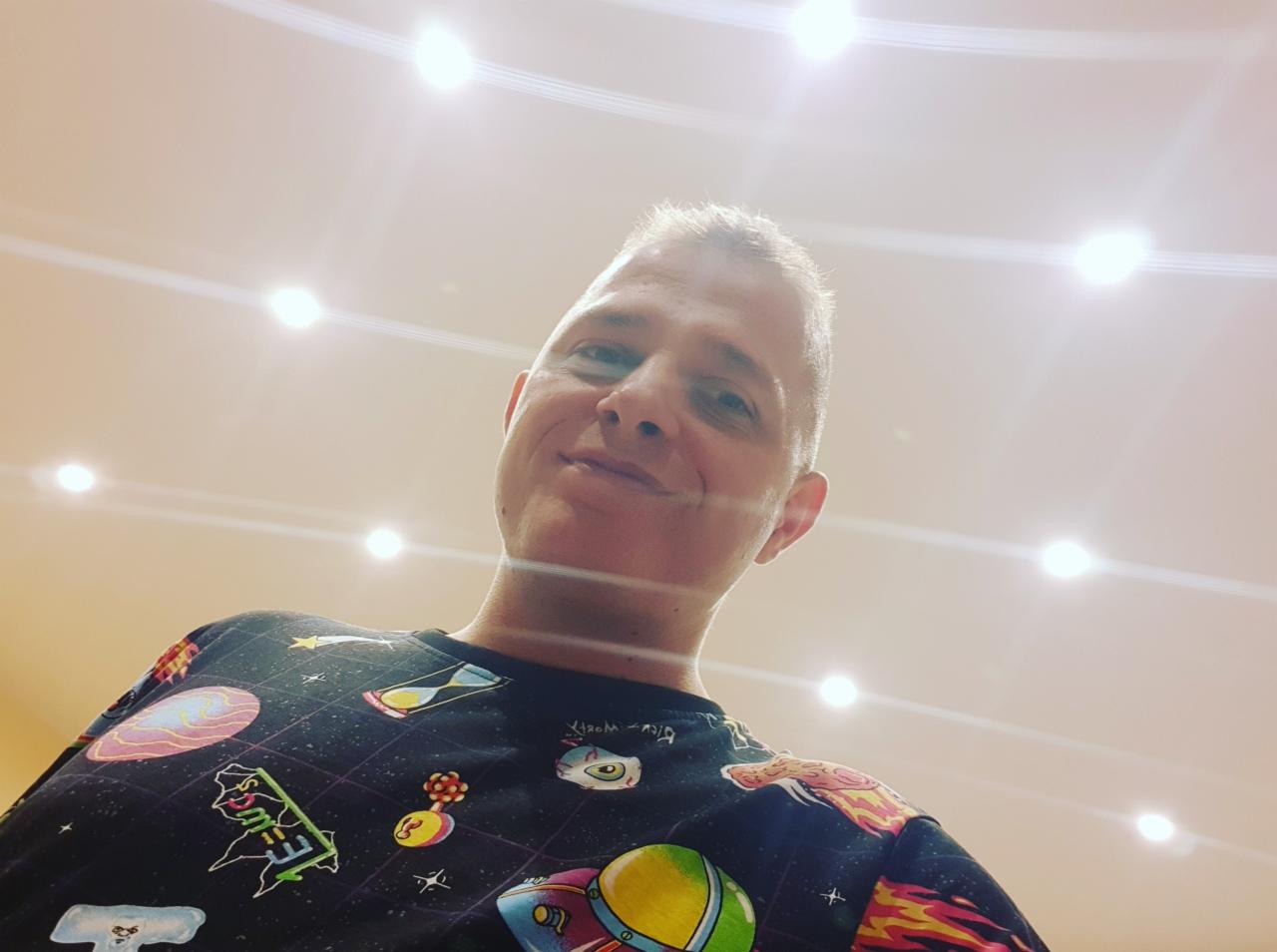 Uśmiechnięty mężczyzna spogląda w dół, w tle sufit z punktowym oświetleniem. Nietypowa perspektywa, koszulka z motywem kosmosu.