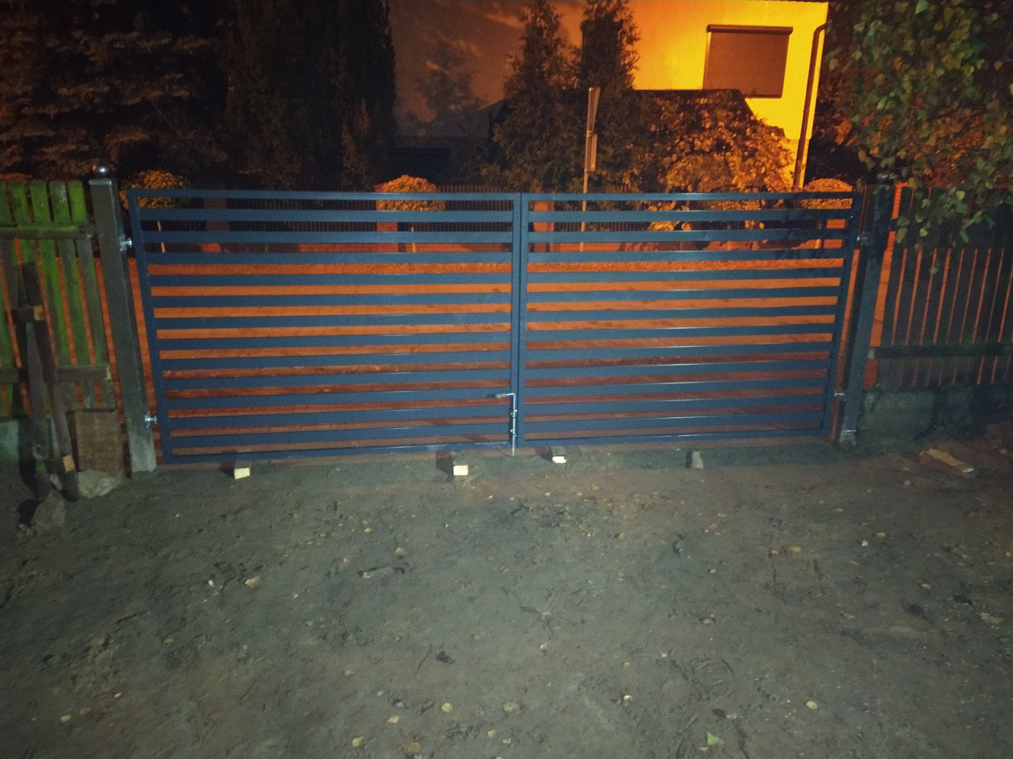 Nowoczesna brama posesyjna z poziomych profili, pomalowana na ciemny kolor, zamontowana między starym, drewnianym płotem a nowym słupkiem, oświetlona światłem zewnętrznym.